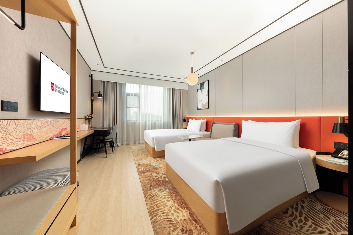 A hotel room with two beds, a desk, a television, and a view of the city.