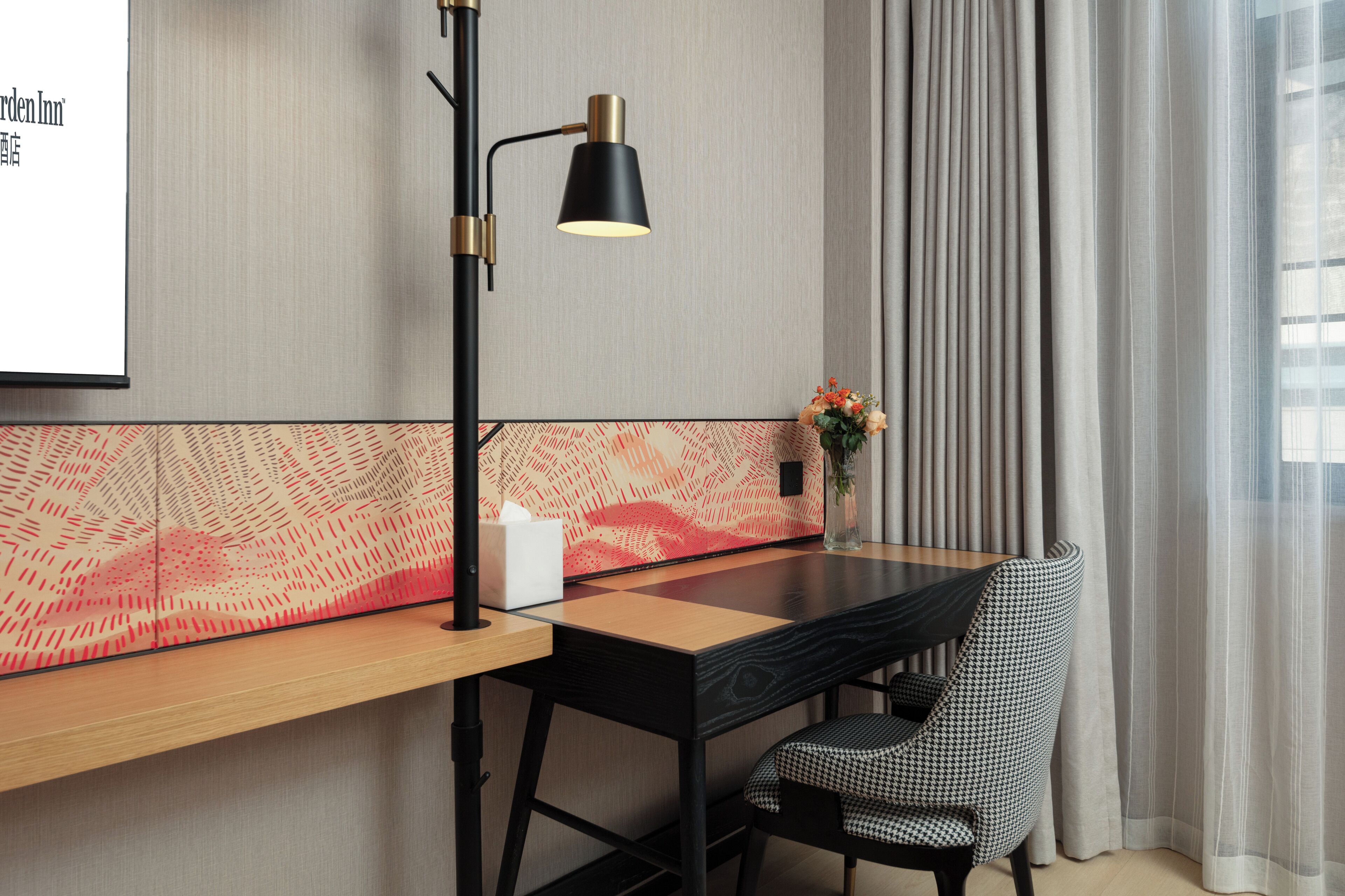 A modern hotel room with a wooden desk, a black lamp, a houndstooth chair, and a decorative wall panel.