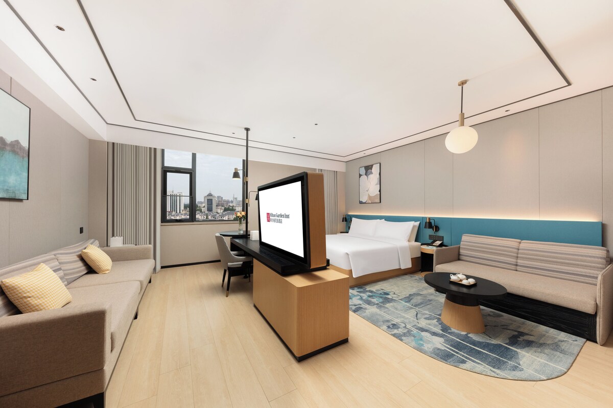 A modern hotel room with a large bed, a sofa, a desk with a flat-screen TV, and a city view through the window.