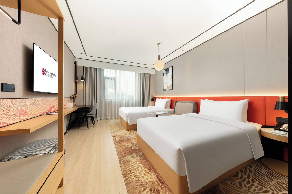 A hotel room with two beds, a desk, a television, and a view of the city.