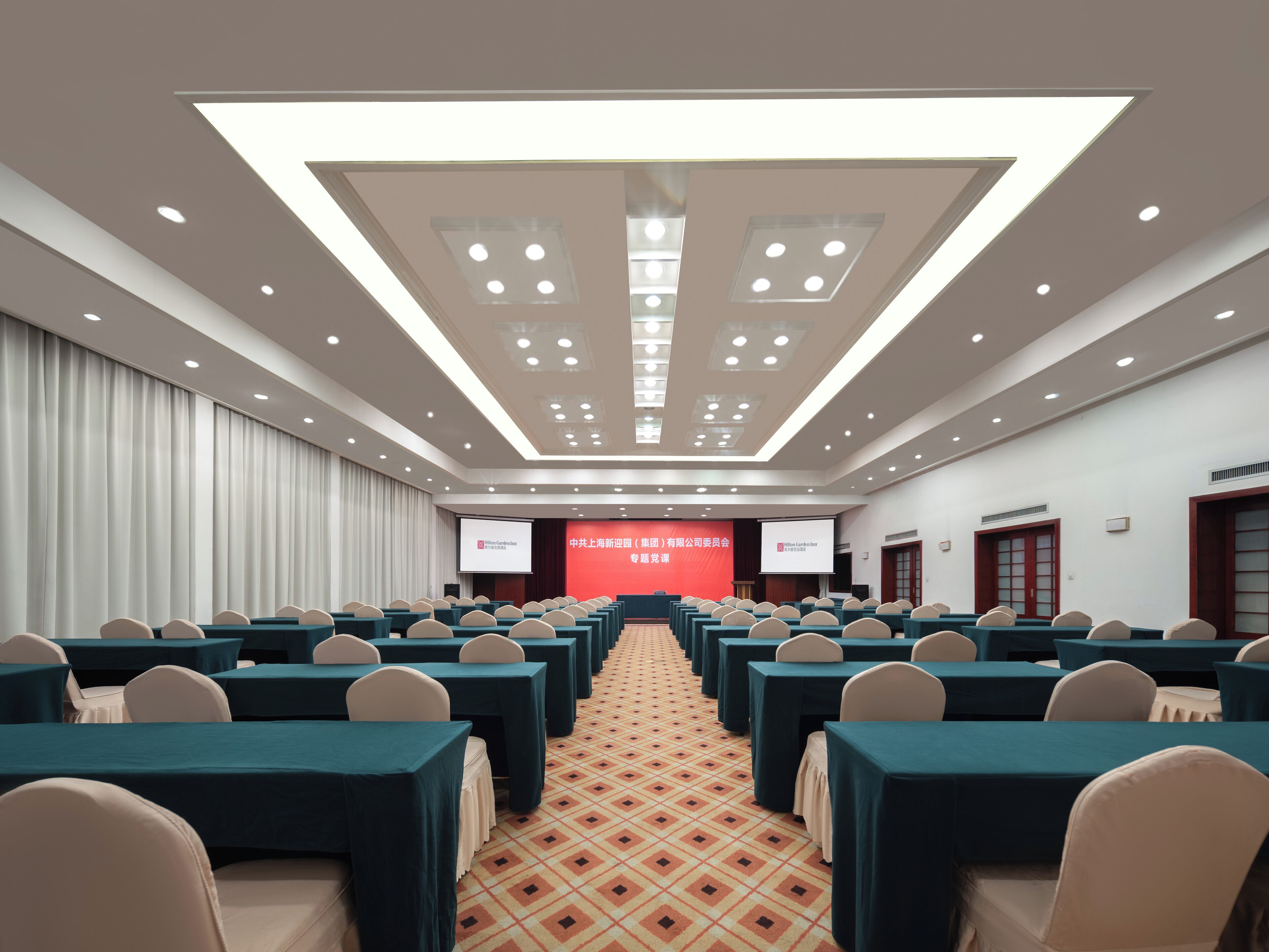 Un salón de conferencias con filas de mesas y sillas, un escenario con un telón de fondo rojo y una alfombra estampada.