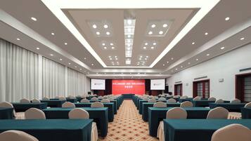 Un salón de conferencias con filas de mesas y sillas, un escenario con un telón de fondo rojo y una alfombra estampada.