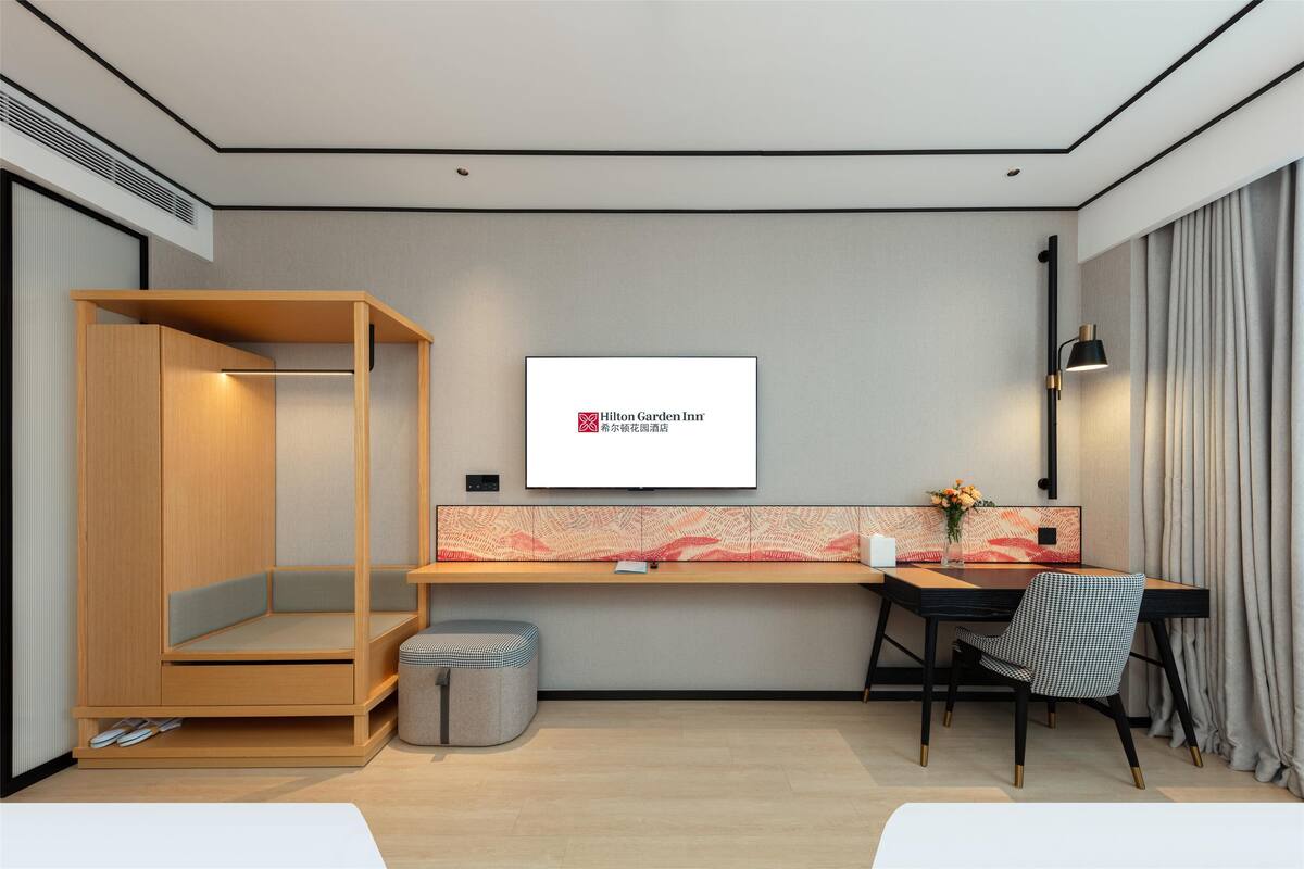A modern hotel room with a wooden wardrobe, a desk with a chair, a small stool, and a wall-mounted TV displaying the Hilton Garden Inn logo.