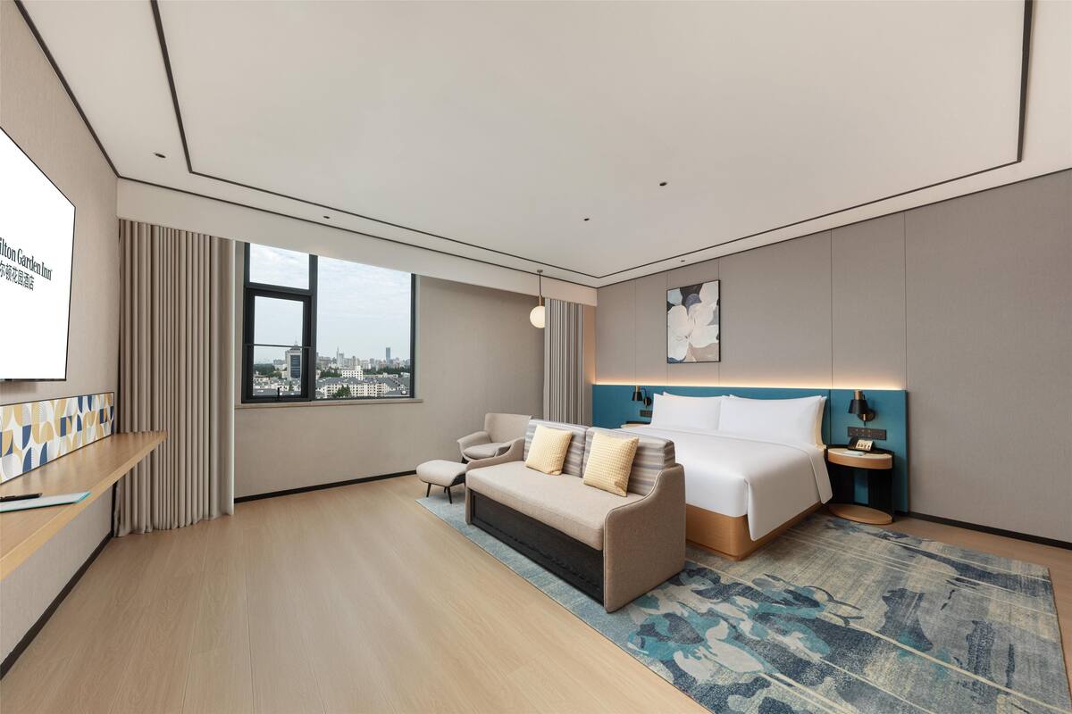 A modern hotel room with a large bed, a sofa, a desk, and a view of the city.