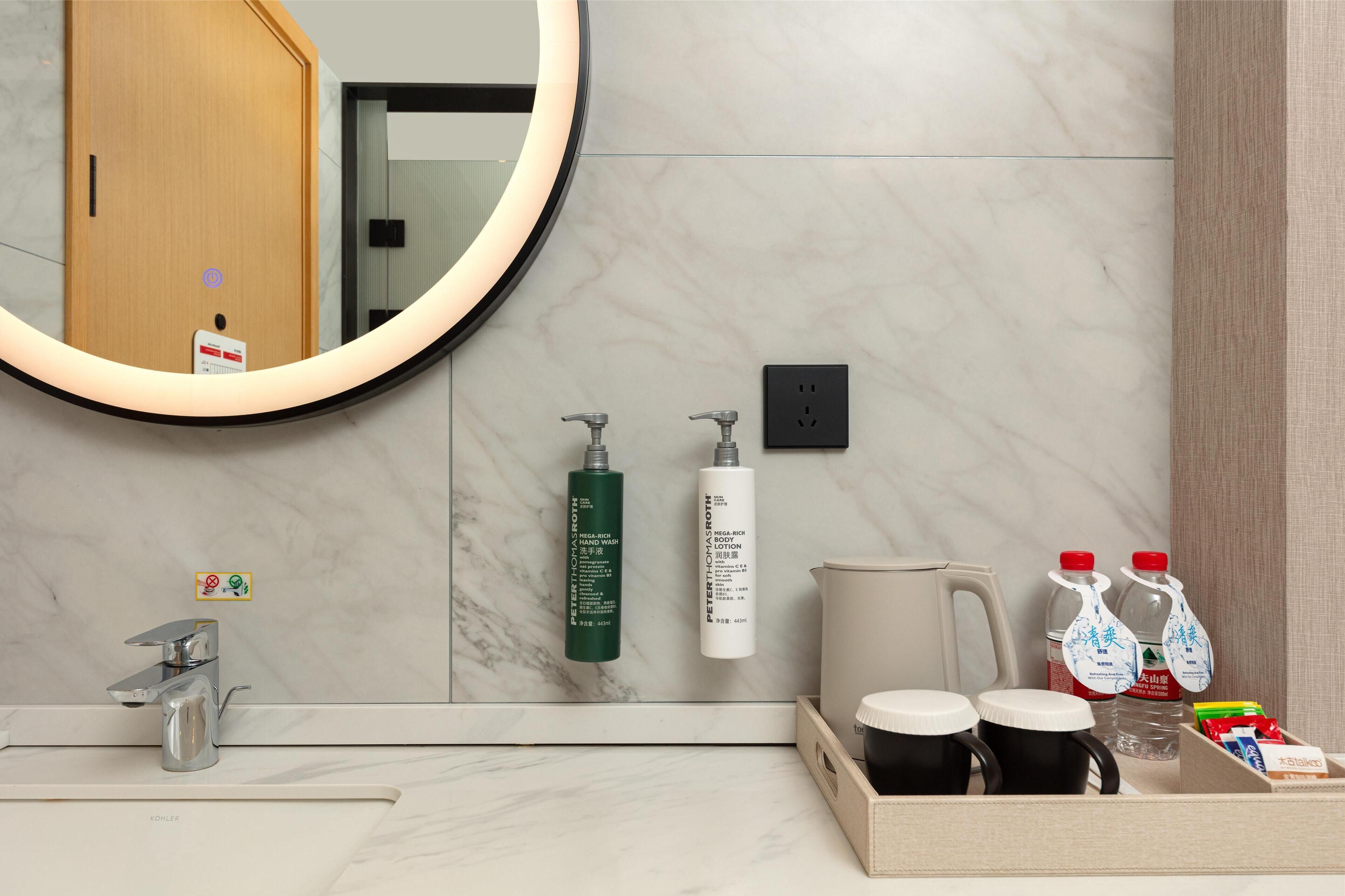 Un baño moderno con un lavamanos de mármol, un espejo redondo, dispensador de jabón para manos, botella de gel de ducha y una cafetera.