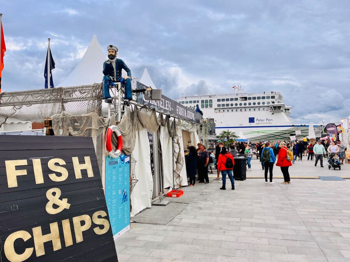 A pier with a sign for 'FISH & CHIPS,' a statue, and a cruise ship in the background.