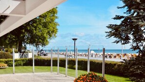 Ein gepflasterter Außenbereich mit Blumenbeet, Blick auf den Strand und einer Umzäunung.