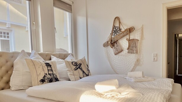 A neatly made bed with white linens and pillows, a hanging bag with a rope handle, and a window with blinds.