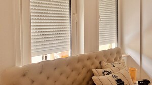 A room with a tufted sofa, two windows with blinds, and a lamp on a side table.