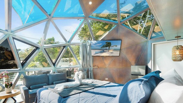 A modern, geometric interior with a large glass ceiling, a bed with blue bedding, a sofa, and a TV on the wall. - Resort-Style Dome - Pool + Hot Tub + 5 Min to Beach (Potrero)
