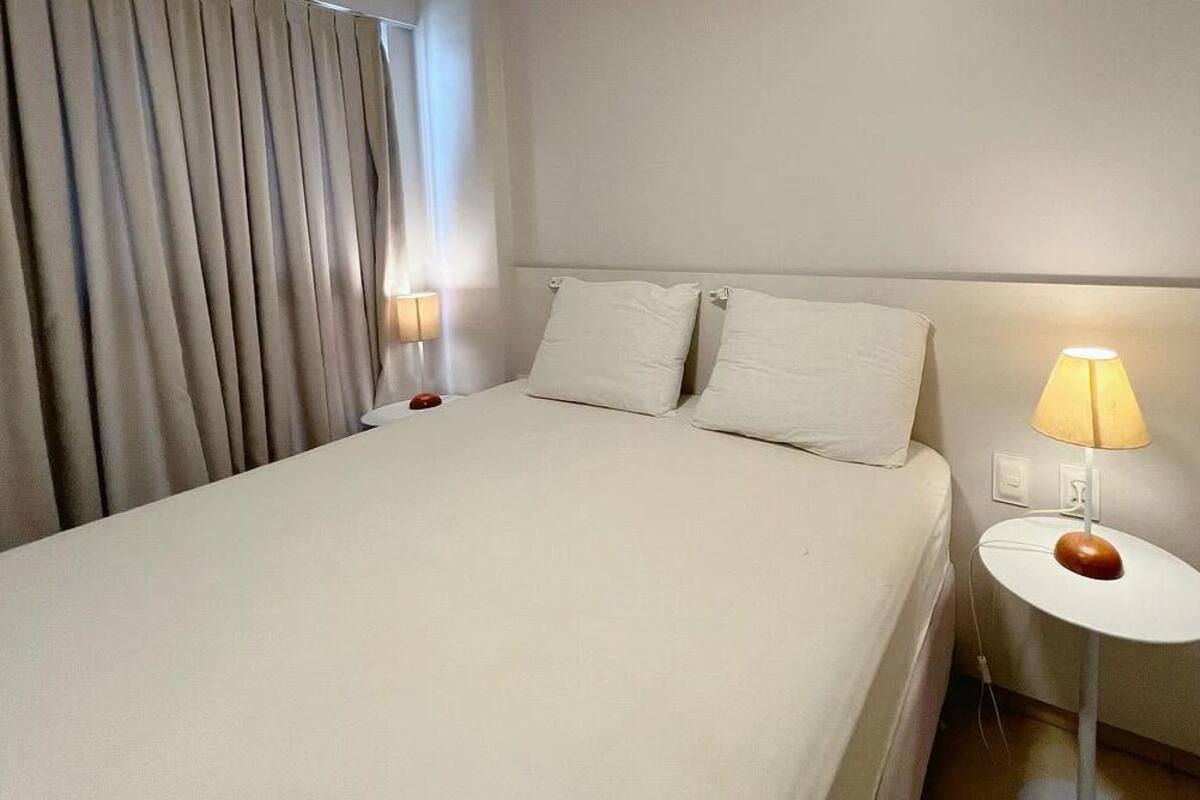 Una habitación de hotel con una cama, dos almohadas, una lámpara de noche y una cortina.