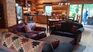 A cozy cabin interior with a living room featuring two sofas, a coffee table, and a fireplace. The kitchen area is visible with wooden cabinets and a refrigerator.