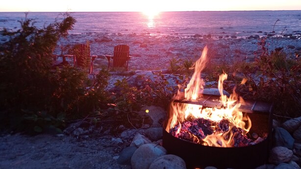 A campfire with rocks and a chair, set against a sunset over the ocean.