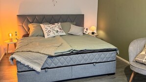 A bedroom with a large bed, a nightstand, a chair, and a wall-mounted light fixture. - Erfurt Lakes vacation apartment - at the gates of the Thuringian state capital  (Nöda)
