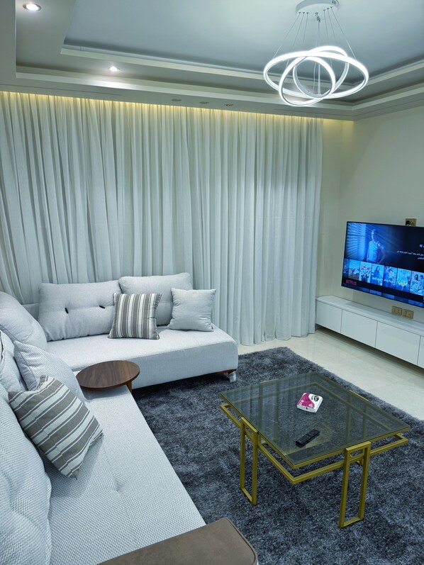 A modern living room with a grey sofa, a glass coffee table, and a flat-screen TV on a white stand.