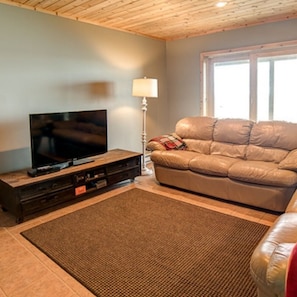 A living room with a television on a wooden stand, a leather sofa, a floor lamp, and a large window. - Beaver Island - Cleveland's Big Chill (Beaver Island)
