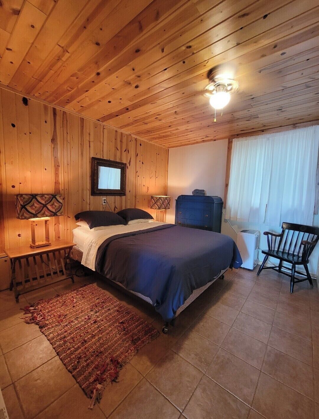 Ein Schlafzimmer mit Holzwänden, einem Bett, Nachttischen, einem Sessel, einem Heizkörper und einem Fenster mit Vorhängen.