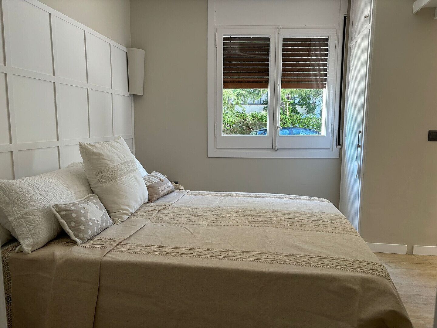 A bedroom with a bed, pillows, a window with blinds, and a view of greenery outside.