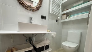 Une salle de bain avec un lavabo blanc, des toilettes et un miroir doté d’un cadre orné. On trouve des paniers de rangement sous le lavabo et des étagères au-dessus des toilettes.