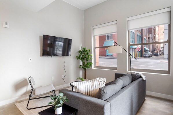 A modern living room with a grey sofa, a small table with a plant, a white chair, and a TV mounted on the wall. - 2 Bedroom Near UC & Good Samaritan Hospital + Wifi (Cincinnati)