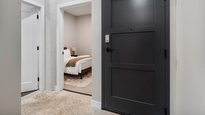 A hallway with a dark door, a bed with a white headboard, and a small bedside table with a lamp. - 2BR Near Fountain Sq.+ Carew Tower Deck | Downtown (Cincinnati)