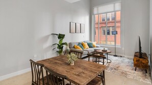 A living room with a wooden dining table, a grey sofa with yellow and teal cushions, a large window with a view of a brick building, and a TV on a wooden stand. - 2BR Near Fountain Sq.+ Carew Tower Deck | Downtown (Cincinnati)
