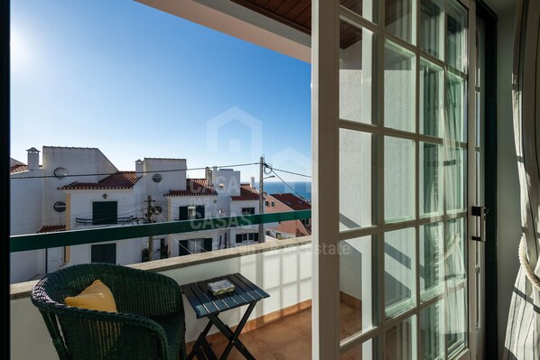 Ein Balkon mit Korbstuhl und kleinem Tisch, mit Blick auf eine Reihe weißer Gebäude mit roten Dächern.