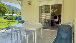 Un patio con una mesa y sillas blancas, un sillón azul y un ventanal corredero que da a un espacio interior con mesa y sillas.