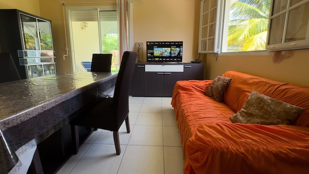 Una habitación con comedor, sofá, televisor y ventana con vista a palmeras.