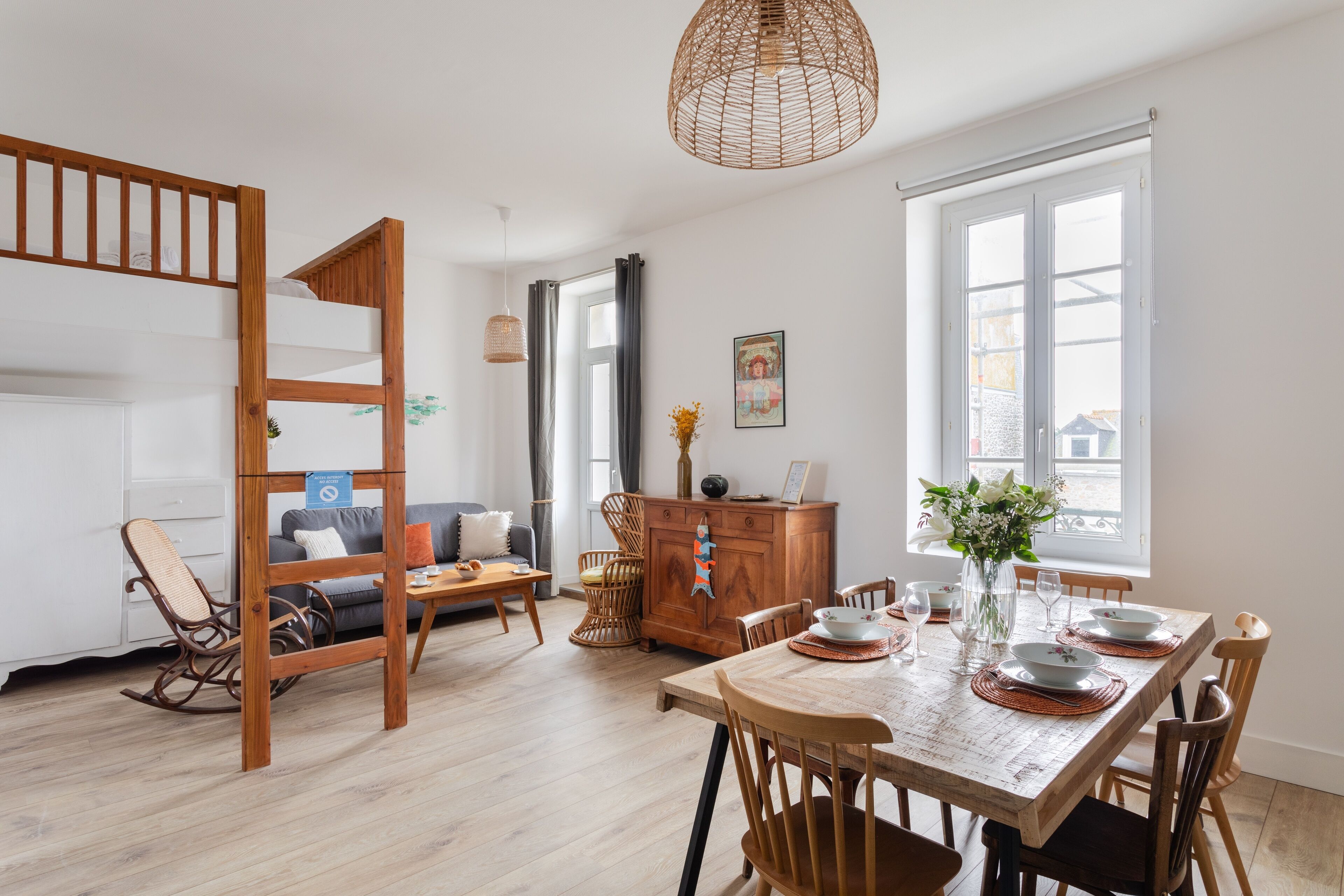 Un salon moderne avec un lit superposé en bois, une table à manger pour quatre personnes et une grande fenêtre laissant entrer la lumière naturelle.
