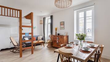 Un salon moderne avec un lit superposé en bois, une table à manger pour quatre personnes et une grande fenêtre laissant entrer la lumière naturelle.