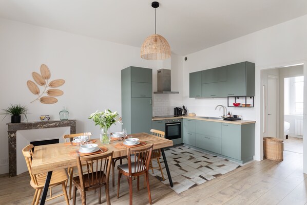 A modern kitchen with teal cabinets, a wooden dining table, and a hanging pendant light. - Coquillages et Crustacés - Apt avec balcon vue mer (Saint-Malo)