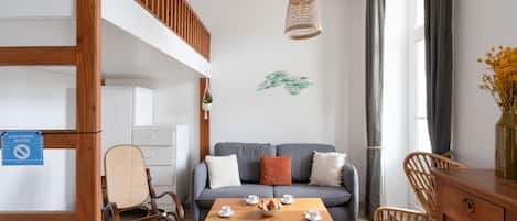 A modern living room with a grey sofa, wooden furniture, a dining table with cups and a bowl, and a hanging light fixture.
