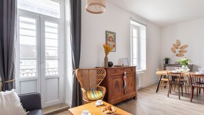 A cozy room with a dining table set for two, a wicker chair, a wooden cabinet, and a window with curtains. - Coquillages et Crustacés - Apt avec balcon vue mer (Saint-Malo)