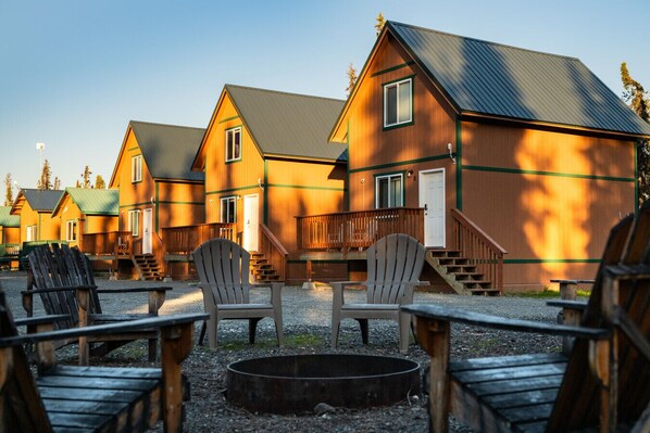 A row of wooden cabins with a fire pit in the center, surrounded by Adirondack chairs.