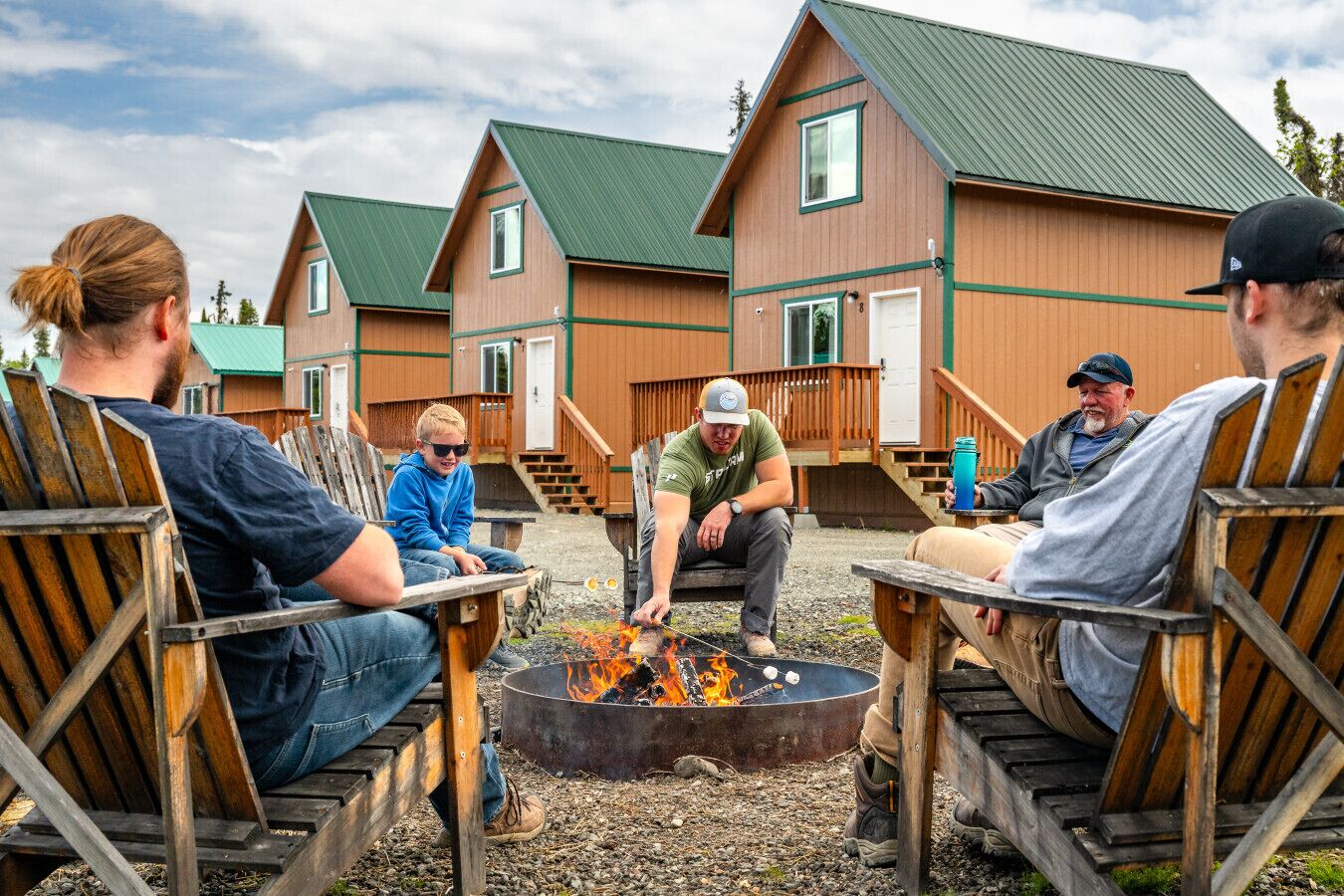A group of people sitting around a campfire in an outdoor setting with wooden cabins in the background.