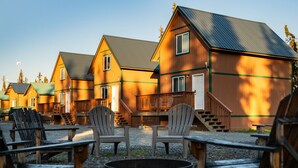A row of wooden cabins with a fire pit in the center, surrounded by Adirondack chairs. - Kenai Fishing Cabin | Walk-In Freezer & Grill (Kenai)