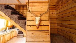 A rustic cabin interior with wooden walls, a kitchen area, and a staircase. - Kenai Fishing Cabin | Walk-In Freezer & Grill (Kenai)
