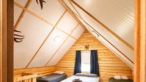 A cozy attic bedroom with a sloped ceiling, wooden walls, a bunk bed, a single bed, a small table, and a window with curtains. - Kenai Fishing Cabin | Walk-In Freezer & Grill (Kenai)
