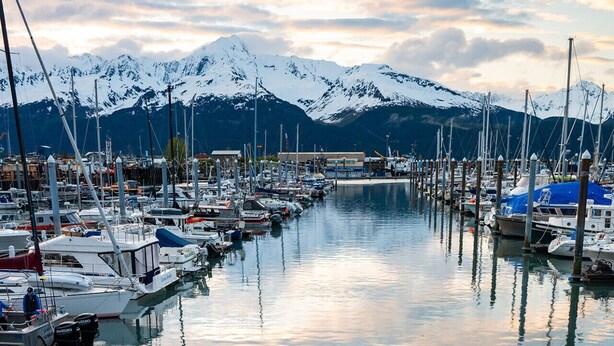 A marina with boats docked, a person walking with a bag, and snow-capped mountains in the background. - Kenai Fishing Cabin | Walk-In Freezer & Grill (Kenai)