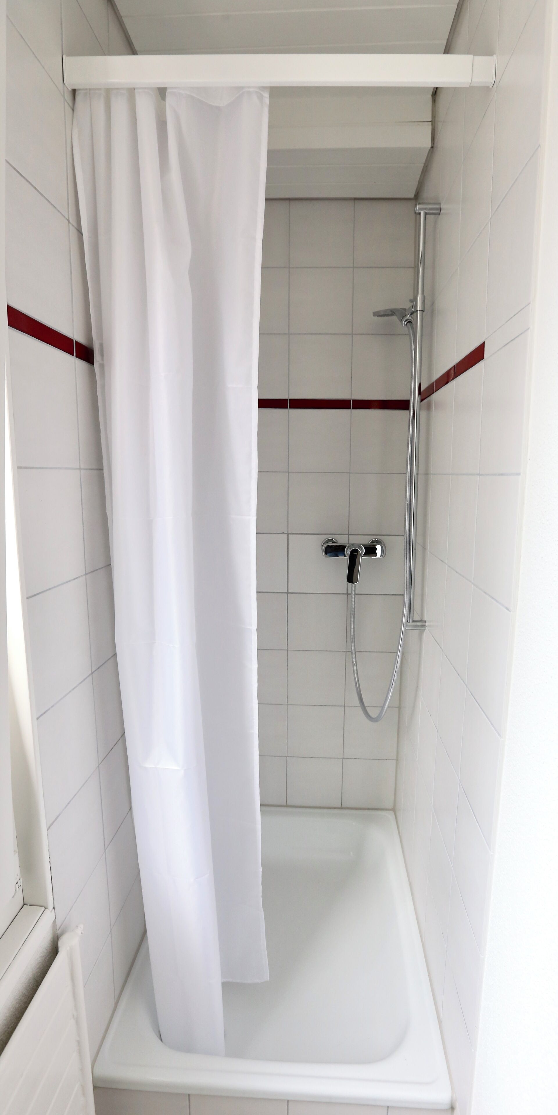 Un espacio de ducha con cortina blanca, un cabezal de ducha de mano y una bañera blanca.