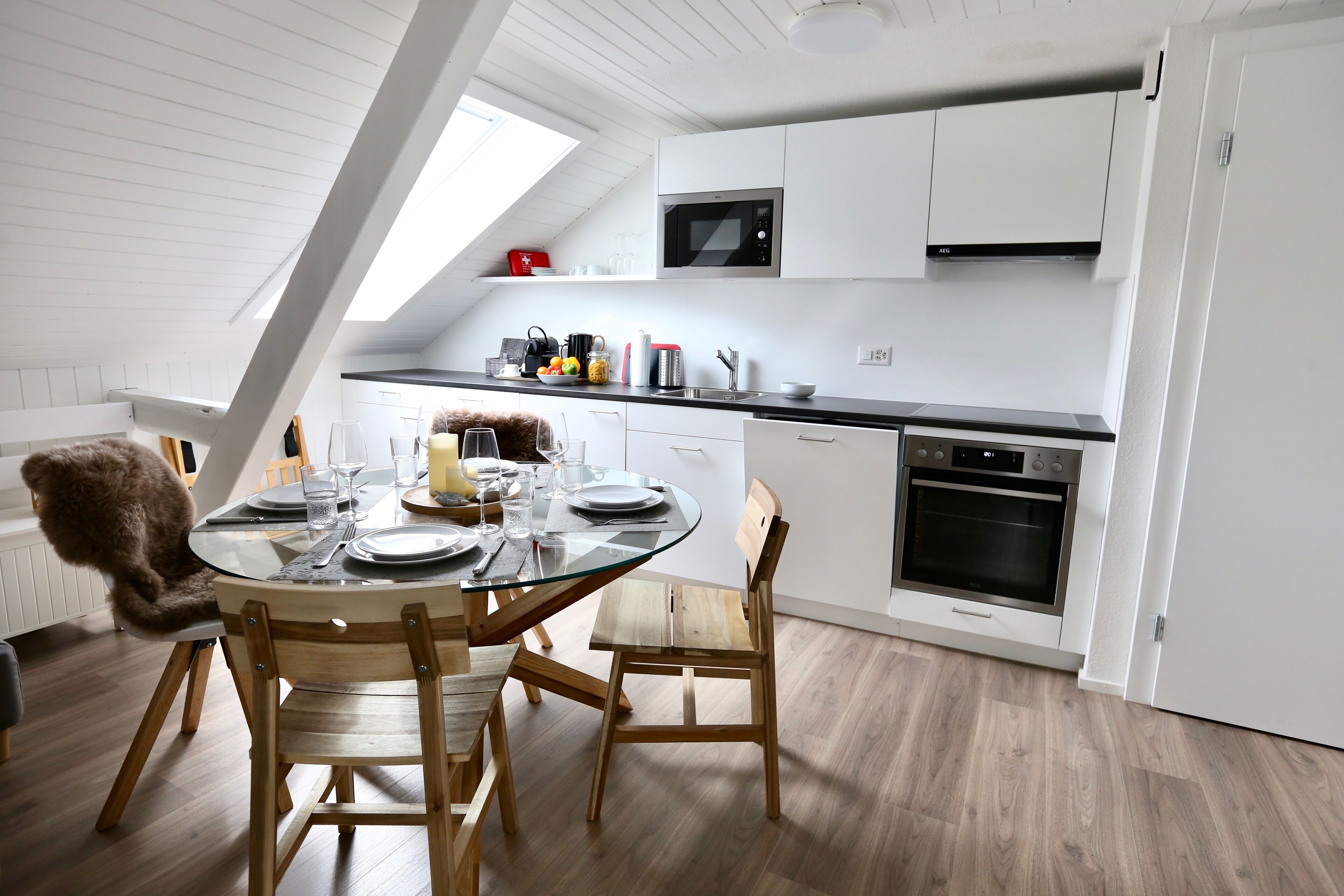 Una cocina moderna con armarios blancos, un microondas y un horno. Hay una mesa de comedor con sillas y una cubierta de vidrio.