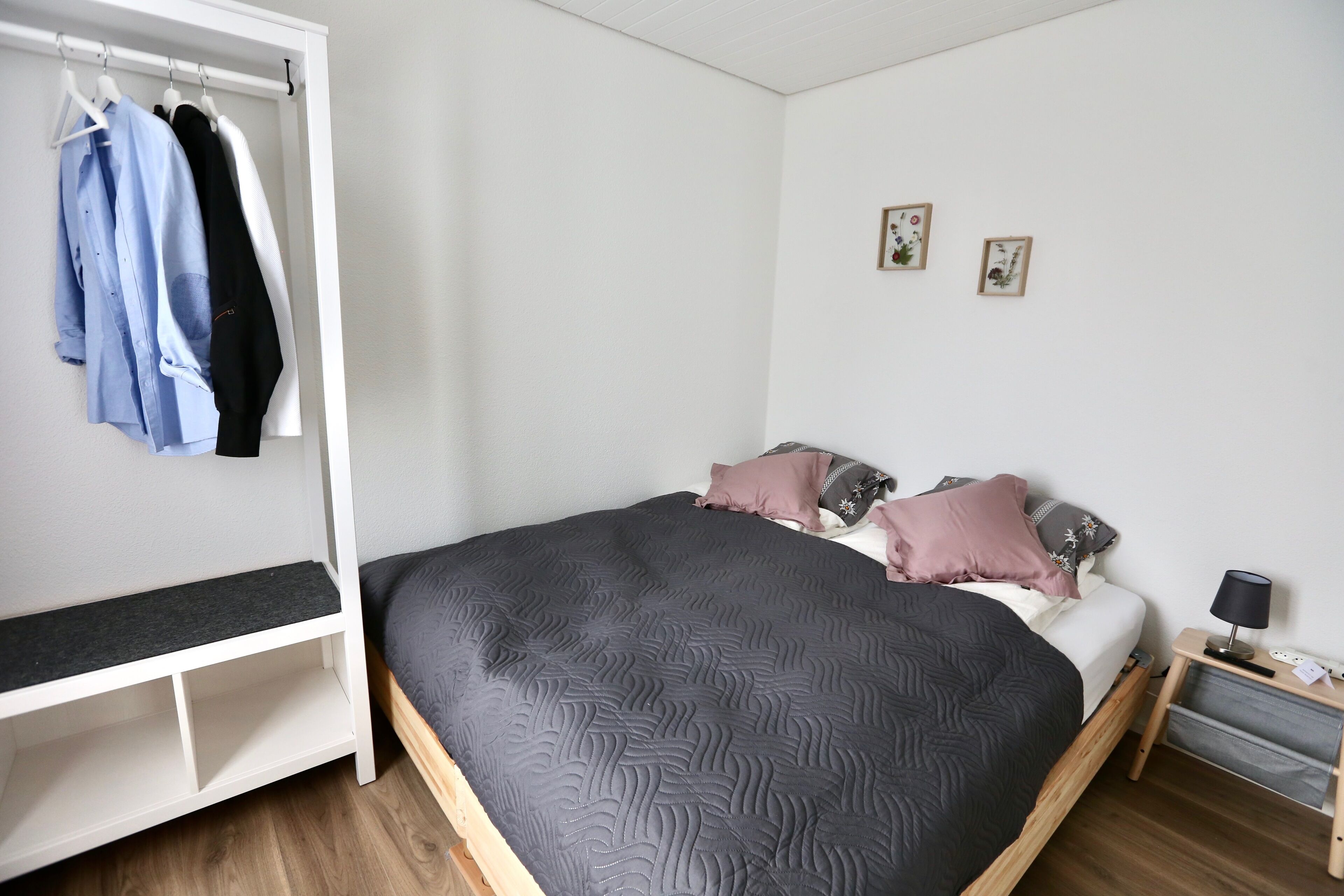 A bedroom with a bed, bedside table, and a wardrobe with hanging clothes.