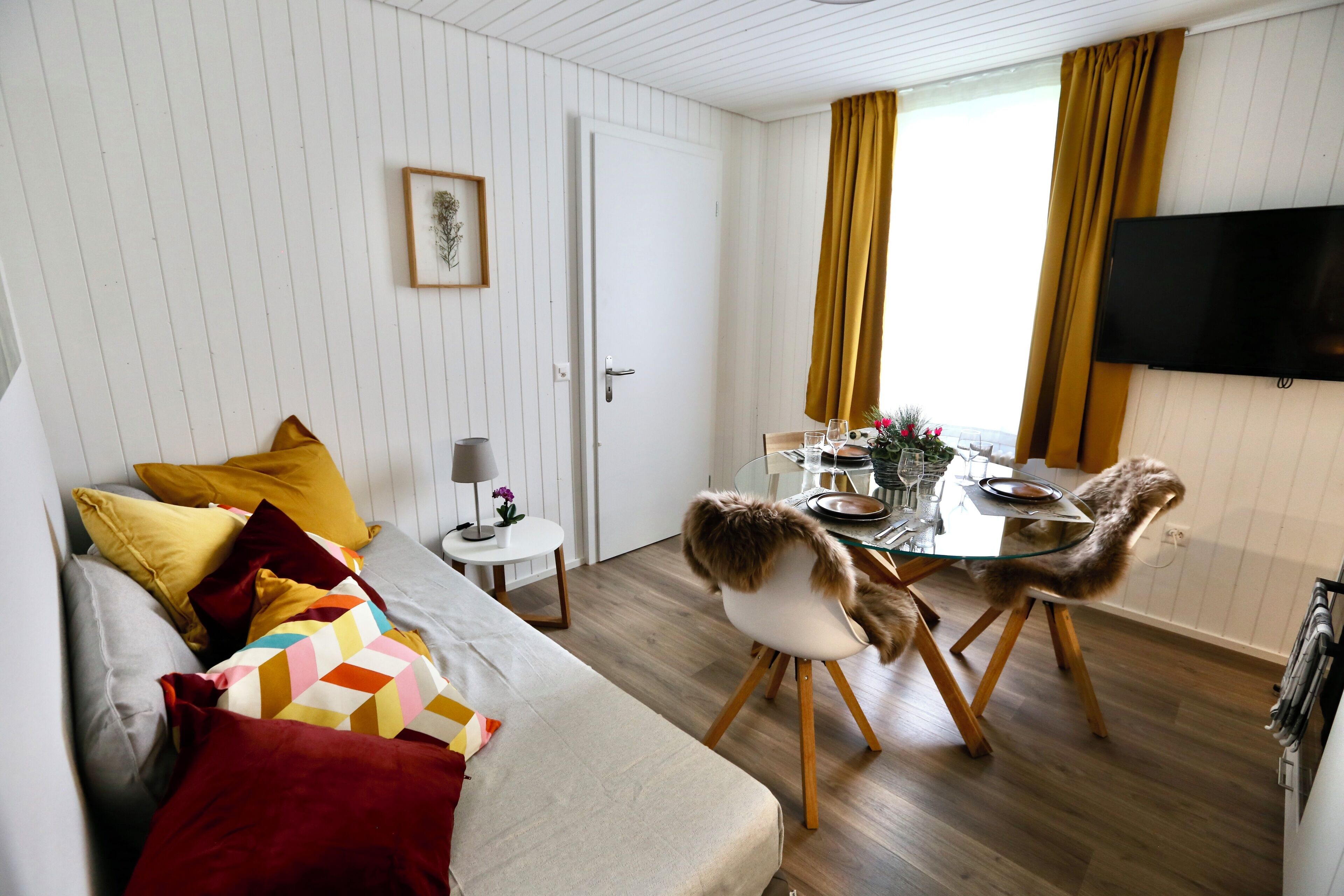 Una sala de estar moderna con un sofá, una mesa de comedor con sillas, un televisor montado en la pared y una puerta.