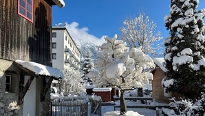 A snow-covered courtyard with a wooden building, a stone wall, and a snow-covered tree. - ALP APARTMENTS with self check-in (Engelberg)
