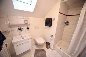 A bathroom with a white sink, toilet, and shower. There is a towel rack with a towel, a trash can, and a shelf with toiletries.