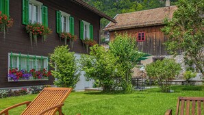 A wooden house with green shutters, a garden with flowers, and a small tree. - ALP APARTMENTS with self check-in (Engelberg)