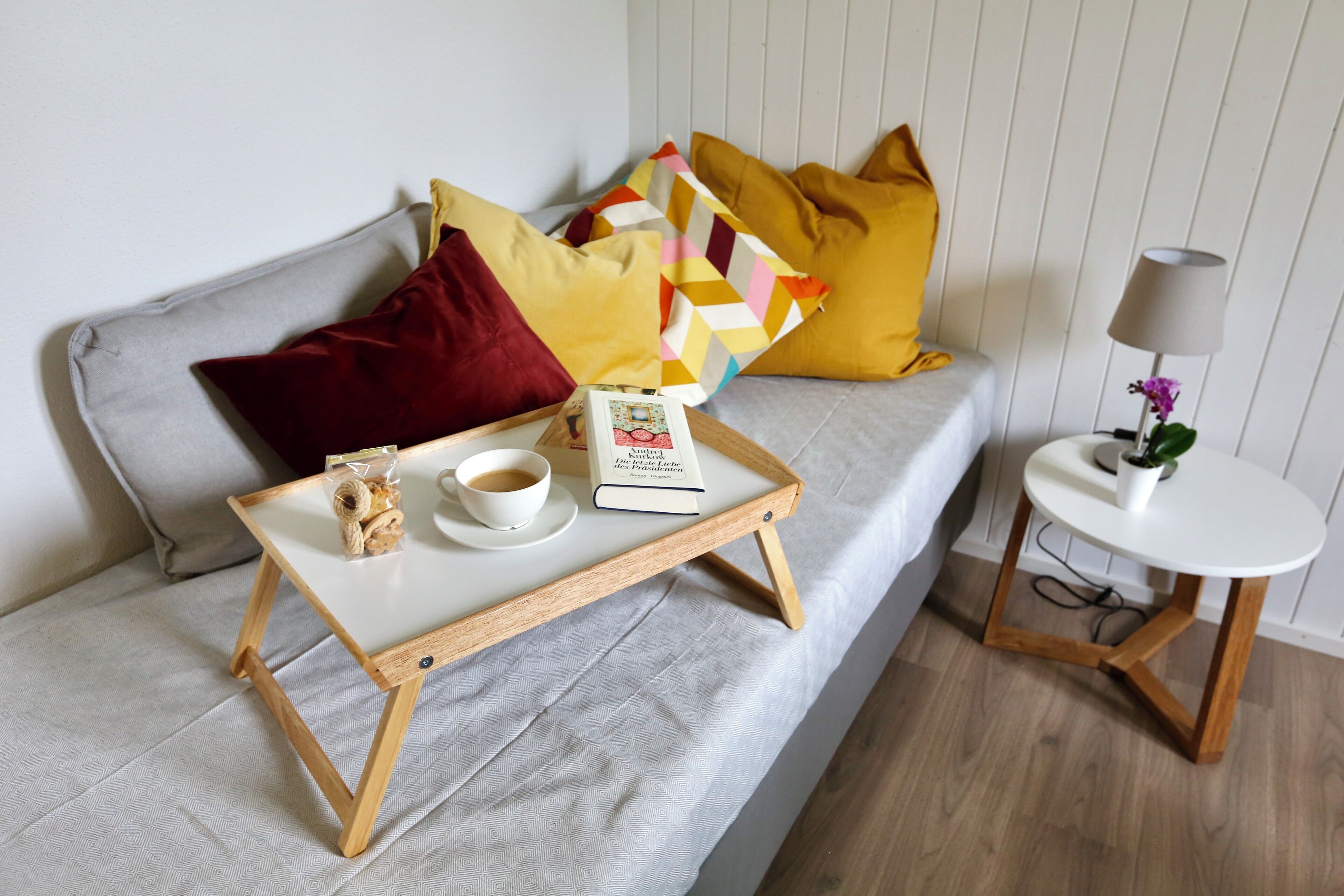 A cozy living area with a grey sofa, a wooden side table with a lamp, and a tray with a book, cup, and snacks.