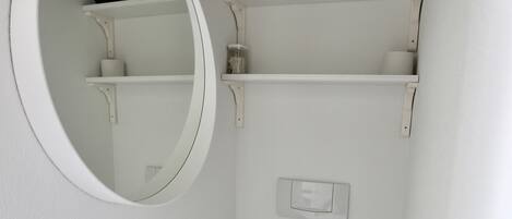 Un baño pequeño y blanco con un inodoro, un espejo y repisas sobre el inodoro.
