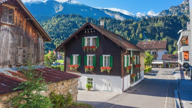 A traditional chalet with a wooden facade, green shutters, and flower boxes. It is located in a mountainous area with a clear sky and snow-capped peaks in the background. - ALP APARTMENTS with self check-in (Engelberg)